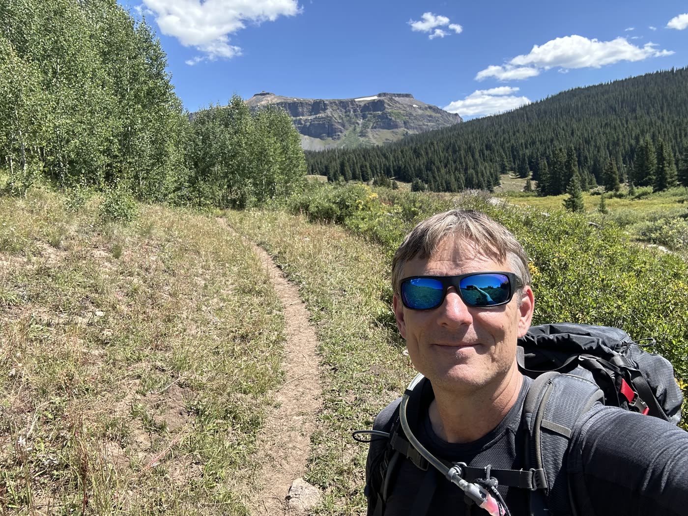 Adrian smiling on a trail with a backpack, mesas and forest behind him
