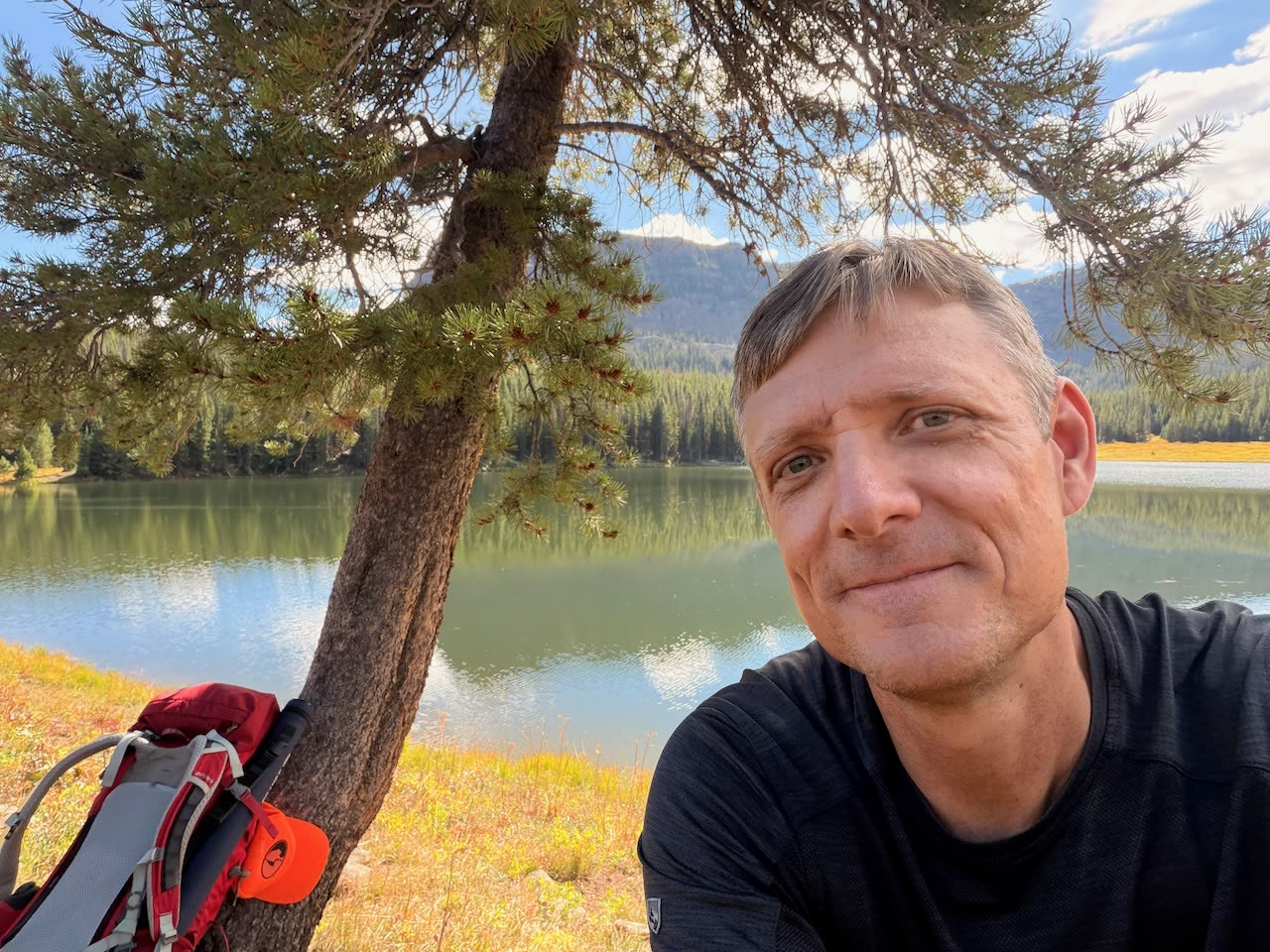 Adrian smiling under a pine on the shore of a mountain lake