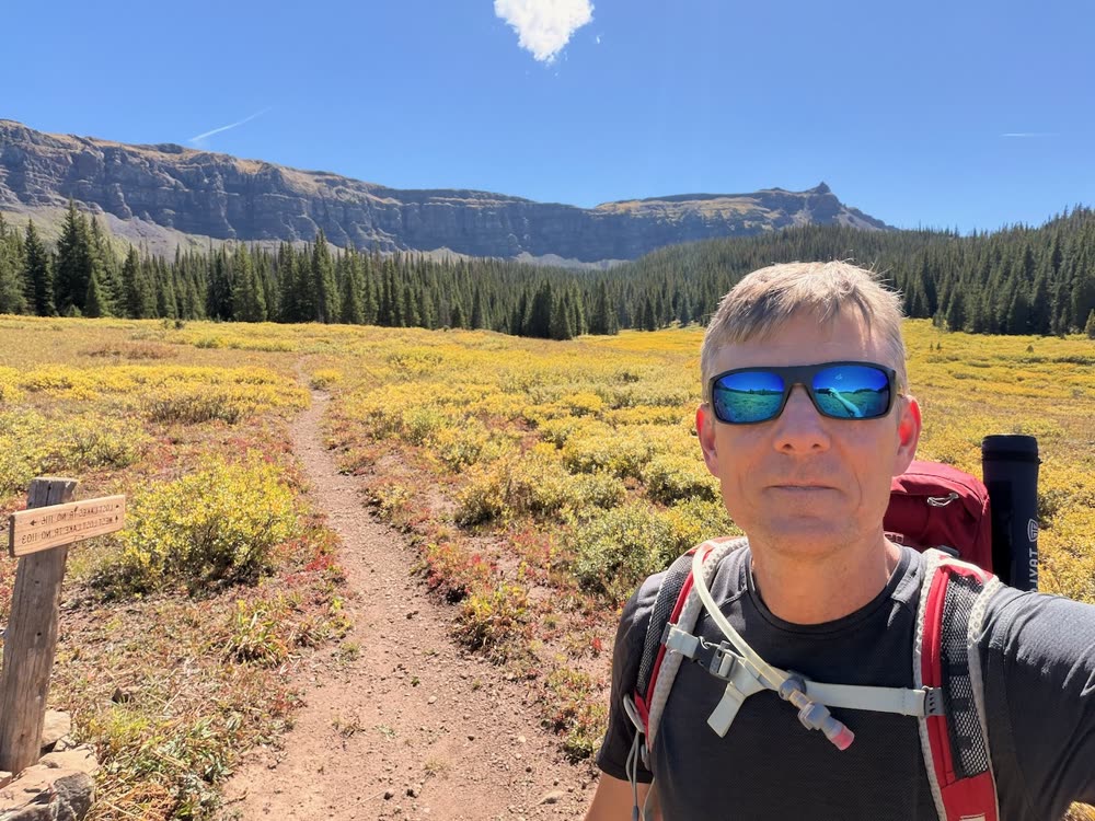 Adrian with a backpack in a wide meadow below a flat-topped mountain