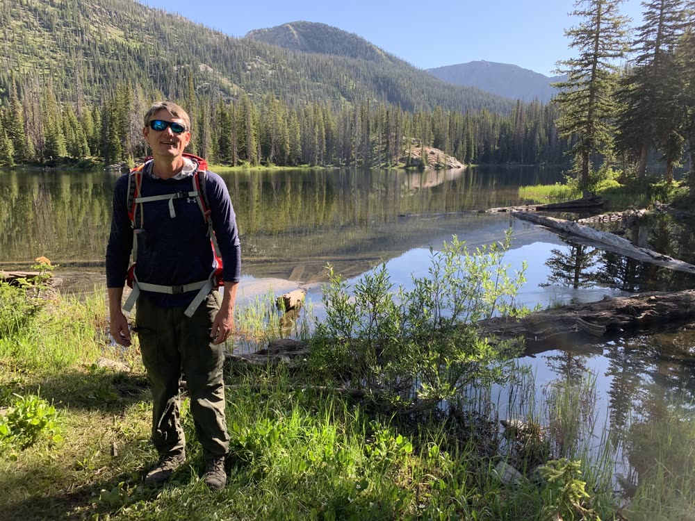 Adrian standing beside a calm alpine lake with a backpack